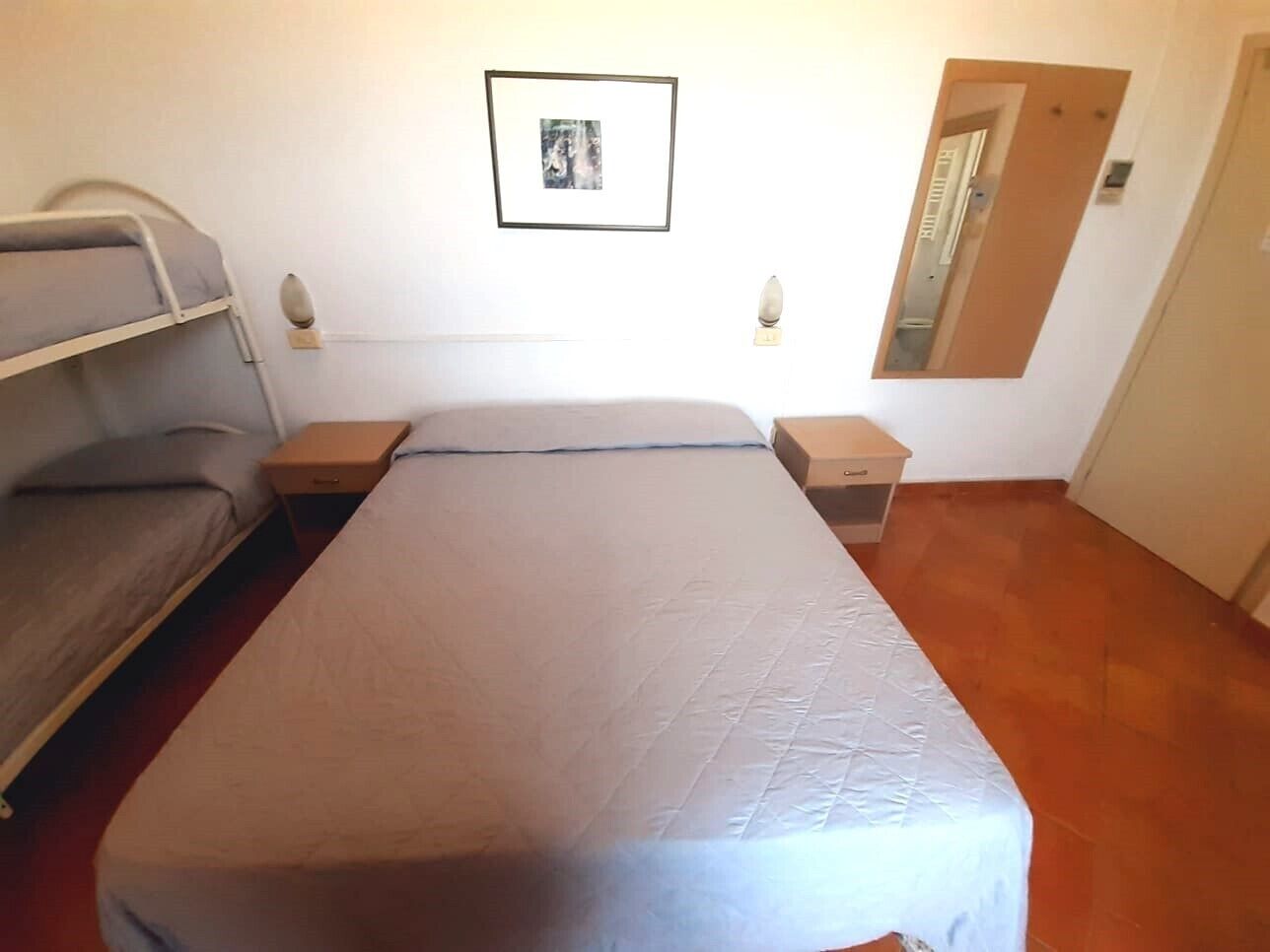 Una cama individual con colcha gris, una mesita de noche y un espejo. También se observa un cuadro colgado en la pared.