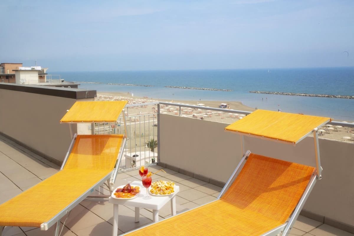 Un balcón con dos tumbonas y una mesa con comida y bebidas, con vista a una playa y al mar.
