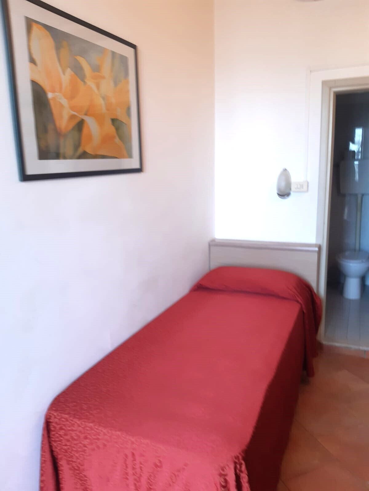 A single bed with a red bedspread in a room with a white wall and a framed painting of flowers.