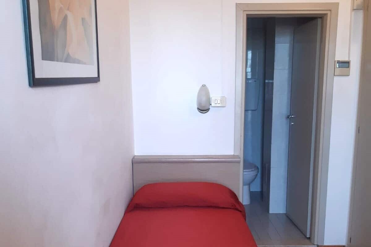 Una habitación pequeña con una cama individual, una colcha roja, un cuadro en la pared y una puerta que da al baño.