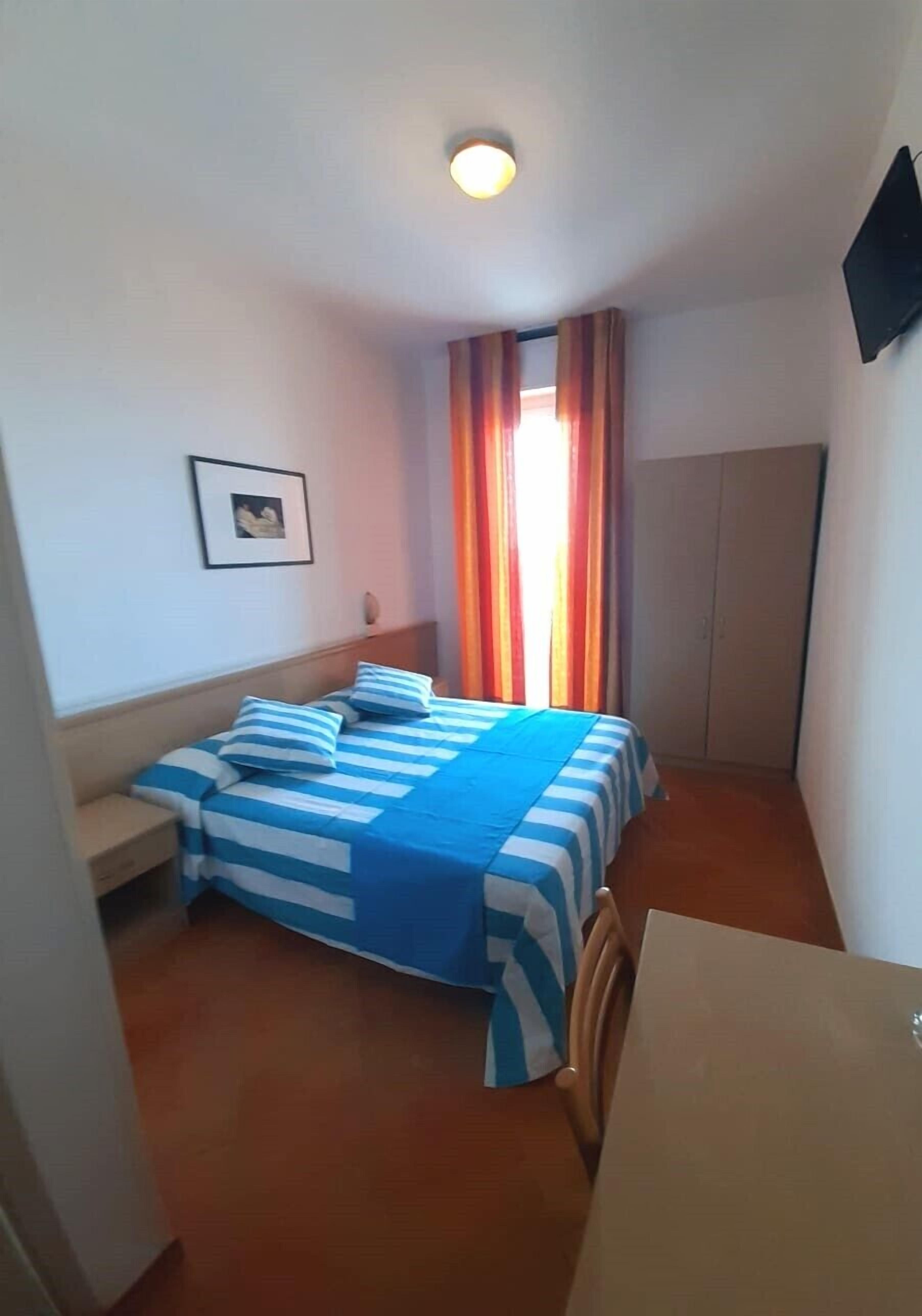 A bedroom with a bed, a nightstand, a desk, a wardrobe, and a window with curtains.