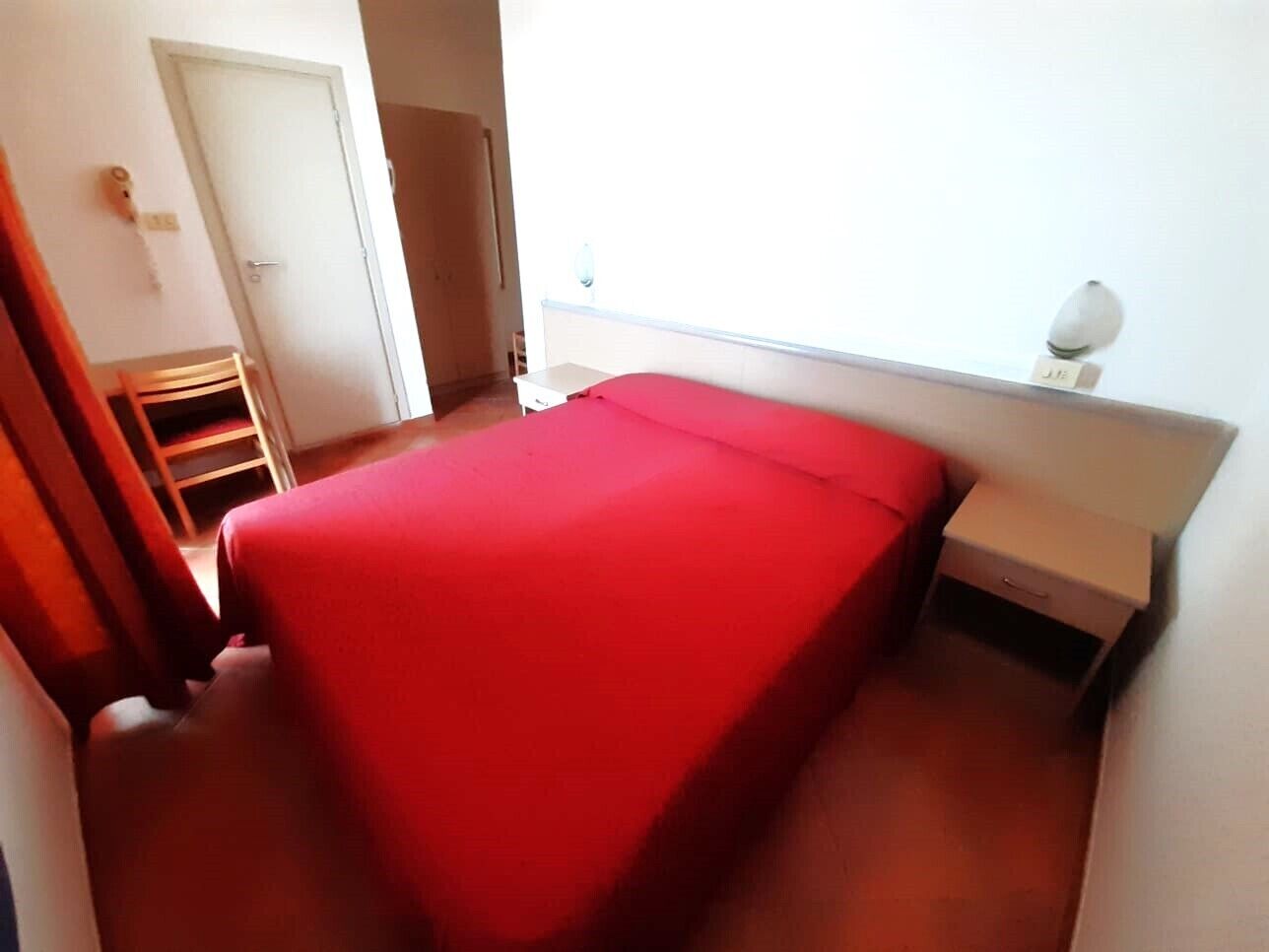 A small, single-bed room with a red bedspread, a bedside table, and a window with a light fixture.