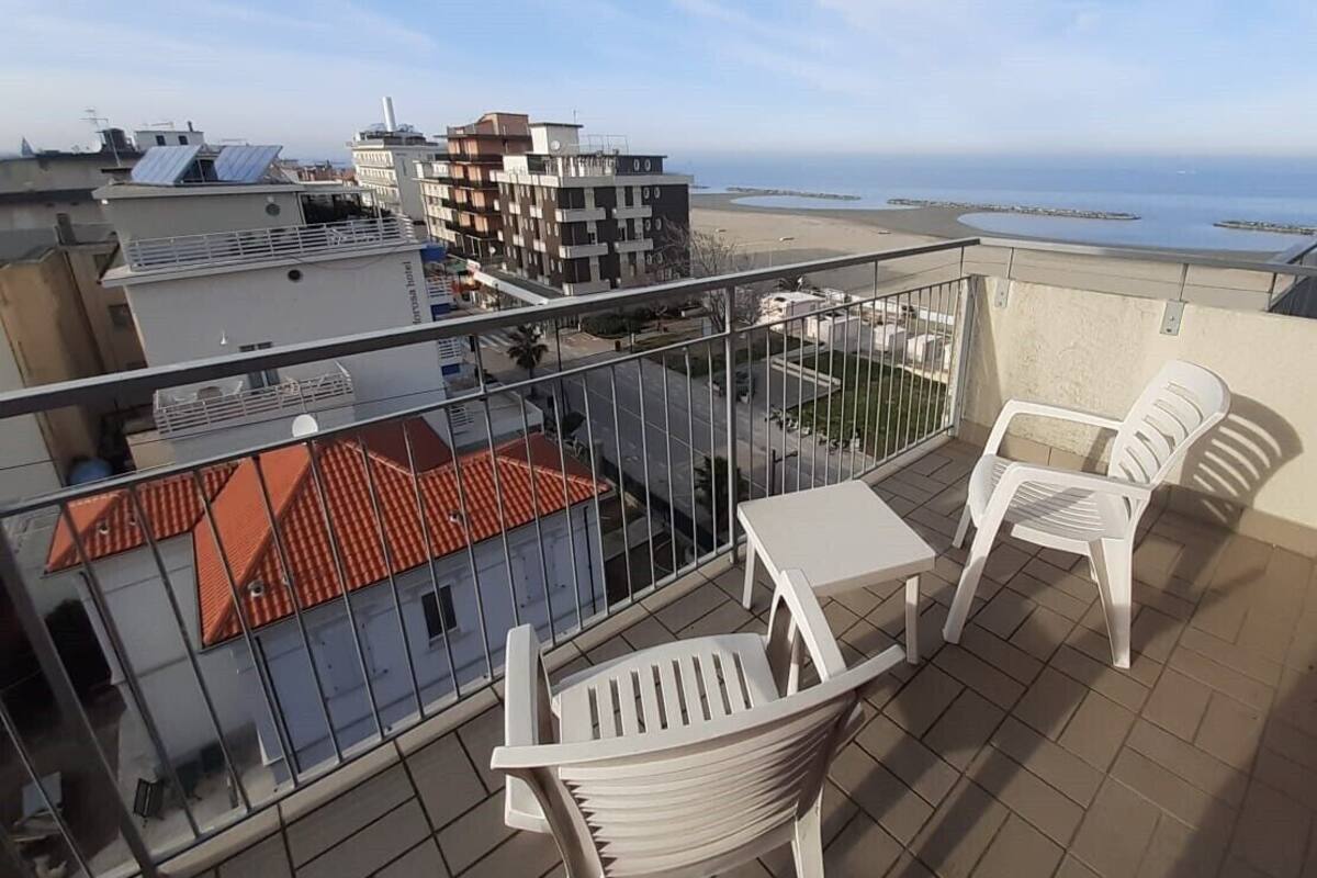 Un balcón con dos sillas de plástico blancas y una mesa pequeña, con vistas a la playa y edificios.