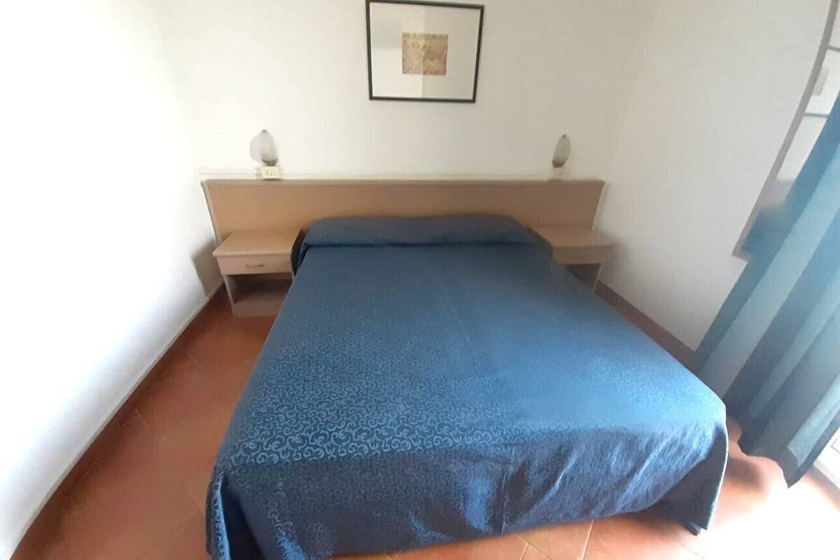 Un dormitorio con cama, mesita de noche, un cuadro enmarcado en la pared y una ventana con cortinas.