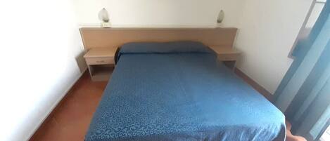 Un dormitorio con cama, mesita de noche, un cuadro enmarcado en la pared y una ventana con cortinas.