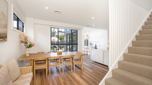 A modern kitchen with a large window, a dining area with a wooden table and chairs, and a staircase with beige carpeted steps. - Burra Beachy - Pet Friendly - 5 Mins Walk to Beach (Culburra Beach)