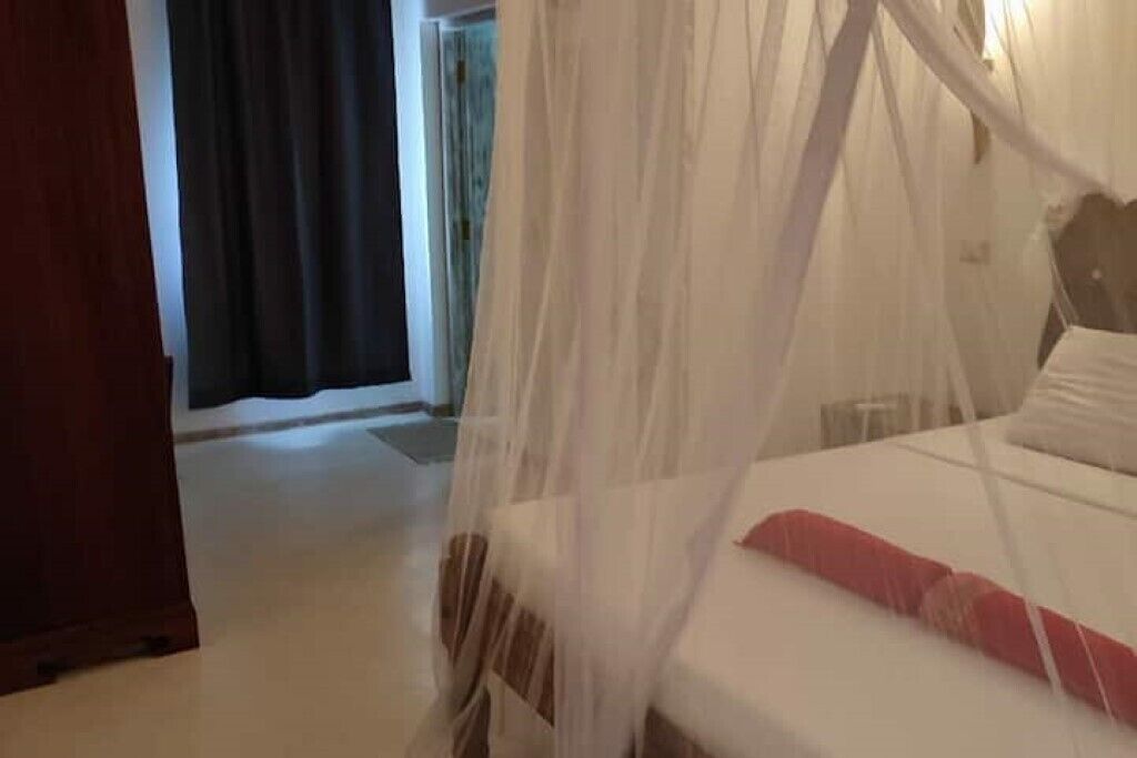 Hotelkamer met een bed onder een witte hemel, een rood kussen en uitzicht op een deur en gordijnen.
