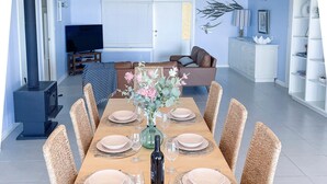 Un coin repas avec une longue table dressée pour six personnes, entourée de chaises en osier. On aperçoit un téléviseur à l’arrière-plan, et une armoire blanche ornée d’objets décoratifs se trouve à droite.