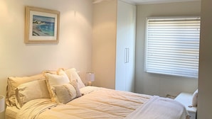 Une chambre à coucher comprenant un lit, une table de chevet, une lampe, un ventilateur de plafond, une fenêtre avec des stores et un tableau encadré accroché au mur.