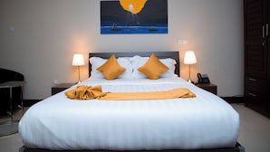 Una cama bien hecha con sábanas blancas y dos almohadas amarillas, un cuadro de un atardecer sobre el agua en la pared, y dos lámparas de noche.