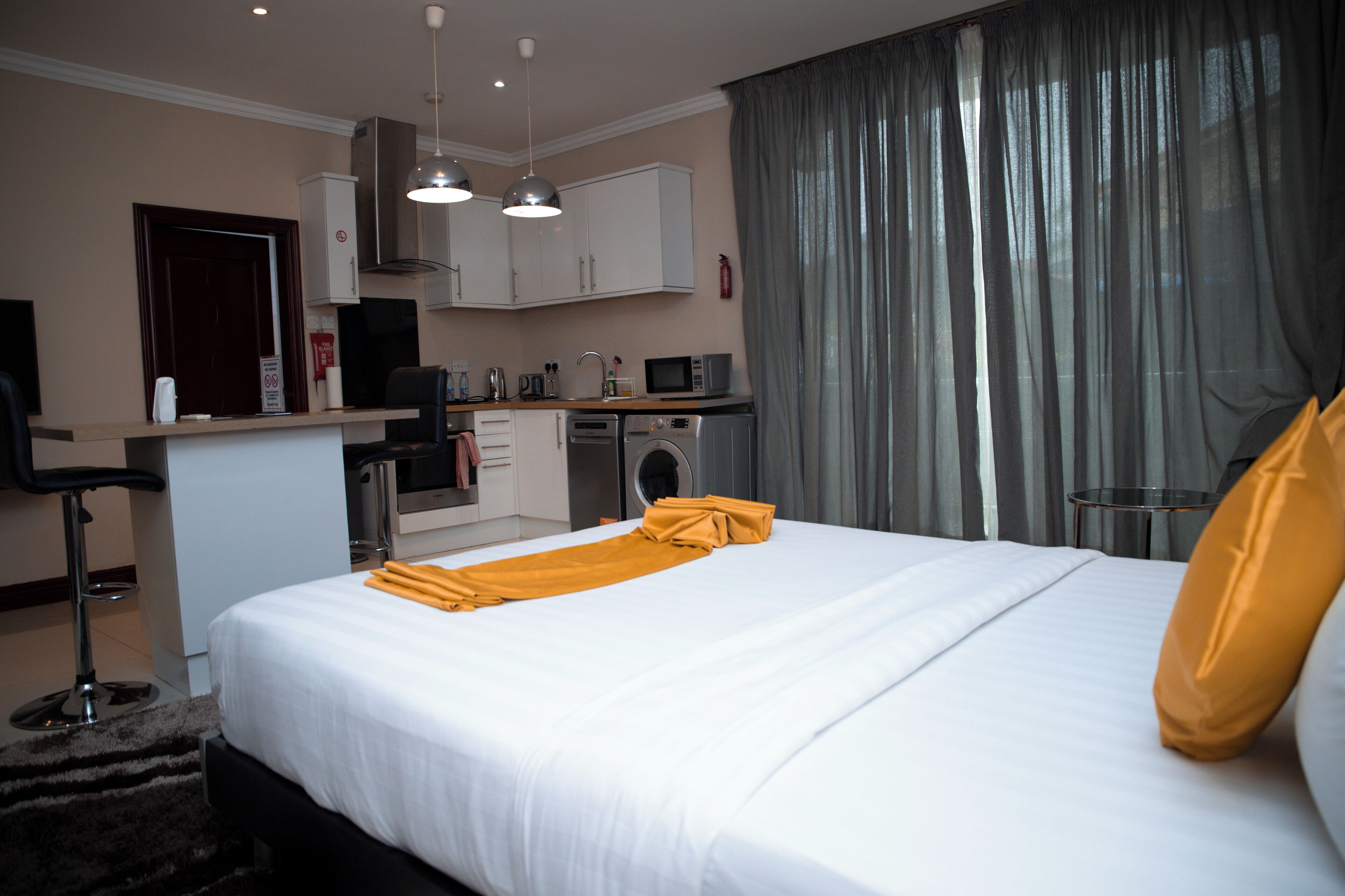 A hotel room with a bed, a desk with a chair, a microwave, a washing machine, and a kitchen area.