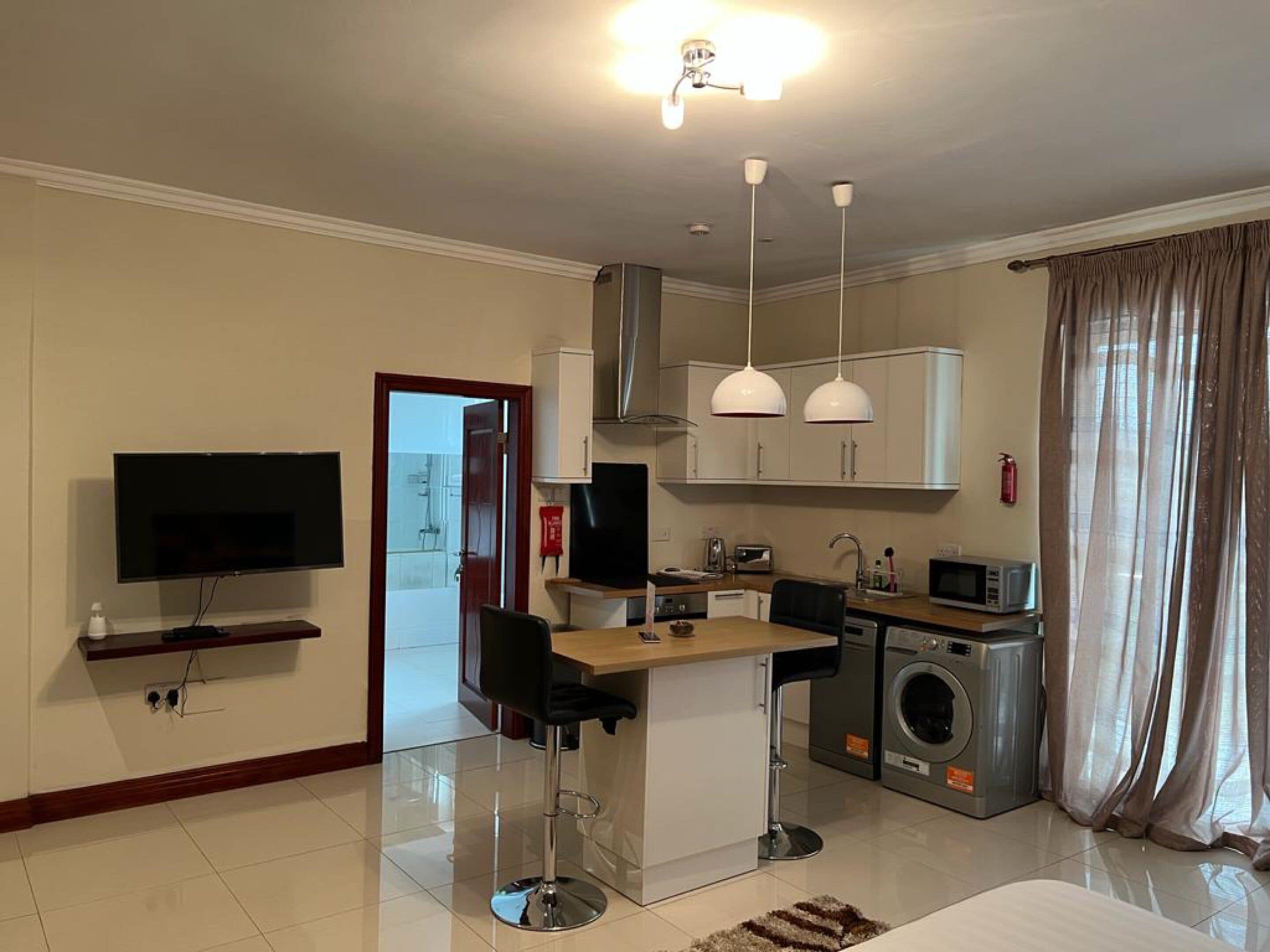 A modern hotel room with a flat-screen TV, a kitchenette with a microwave, a washing machine, and a dining area with a bar stool.