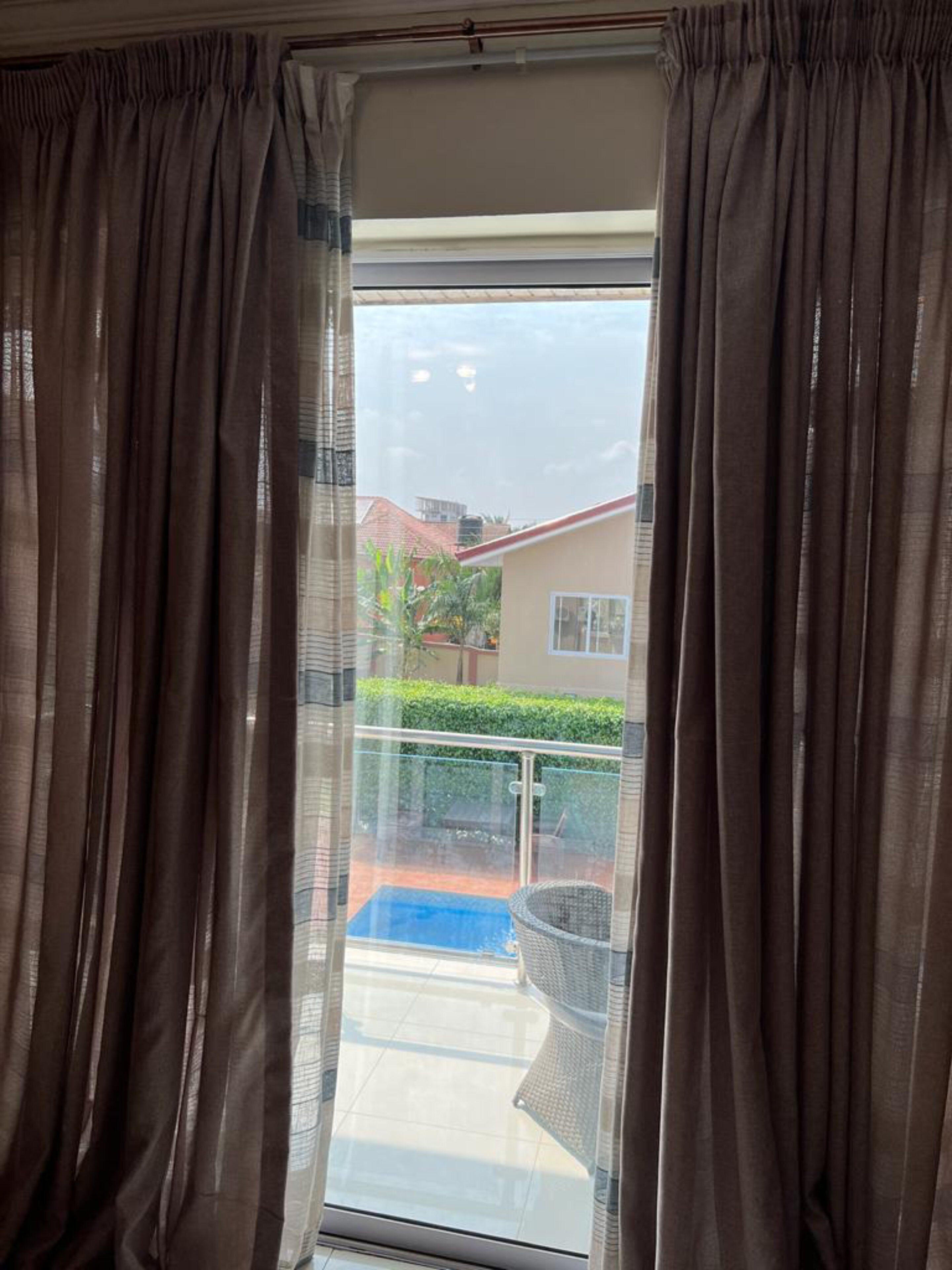 A view through a window with curtains, revealing a balcony with a blue mat, a wicker chair, and a hedge.