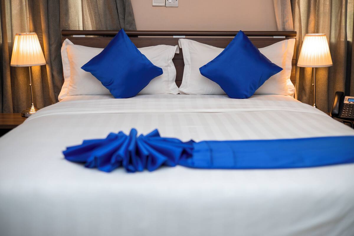 A hotel room with a neatly made bed, two blue pillows, and a blue towel.