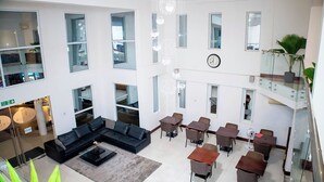 Un amplio hall de hotel de varios niveles con un diseño moderno, que cuenta con una gran zona de estar, varias mesas de comedor y un reloj visible en la pared.
