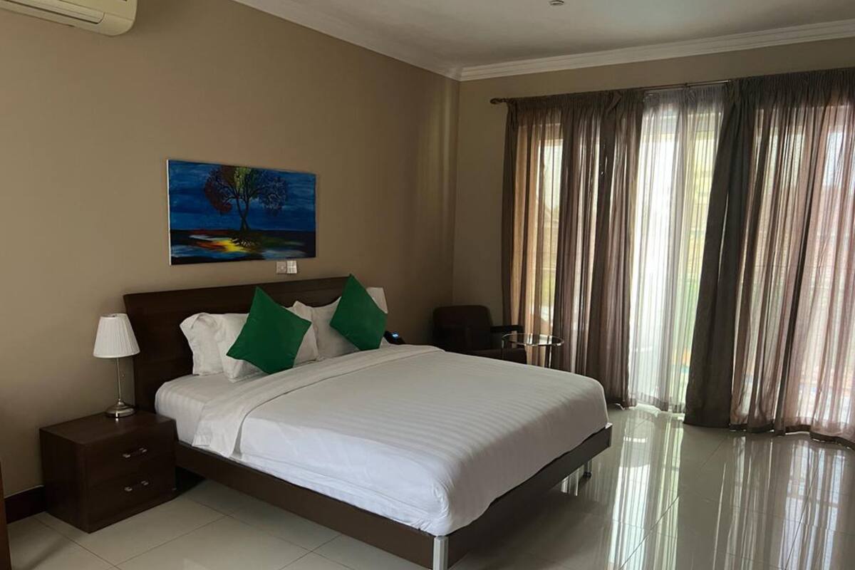 A hotel room with a large bed, a nightstand, a lamp, a painting on the wall, and a view of the outdoors through curtains.