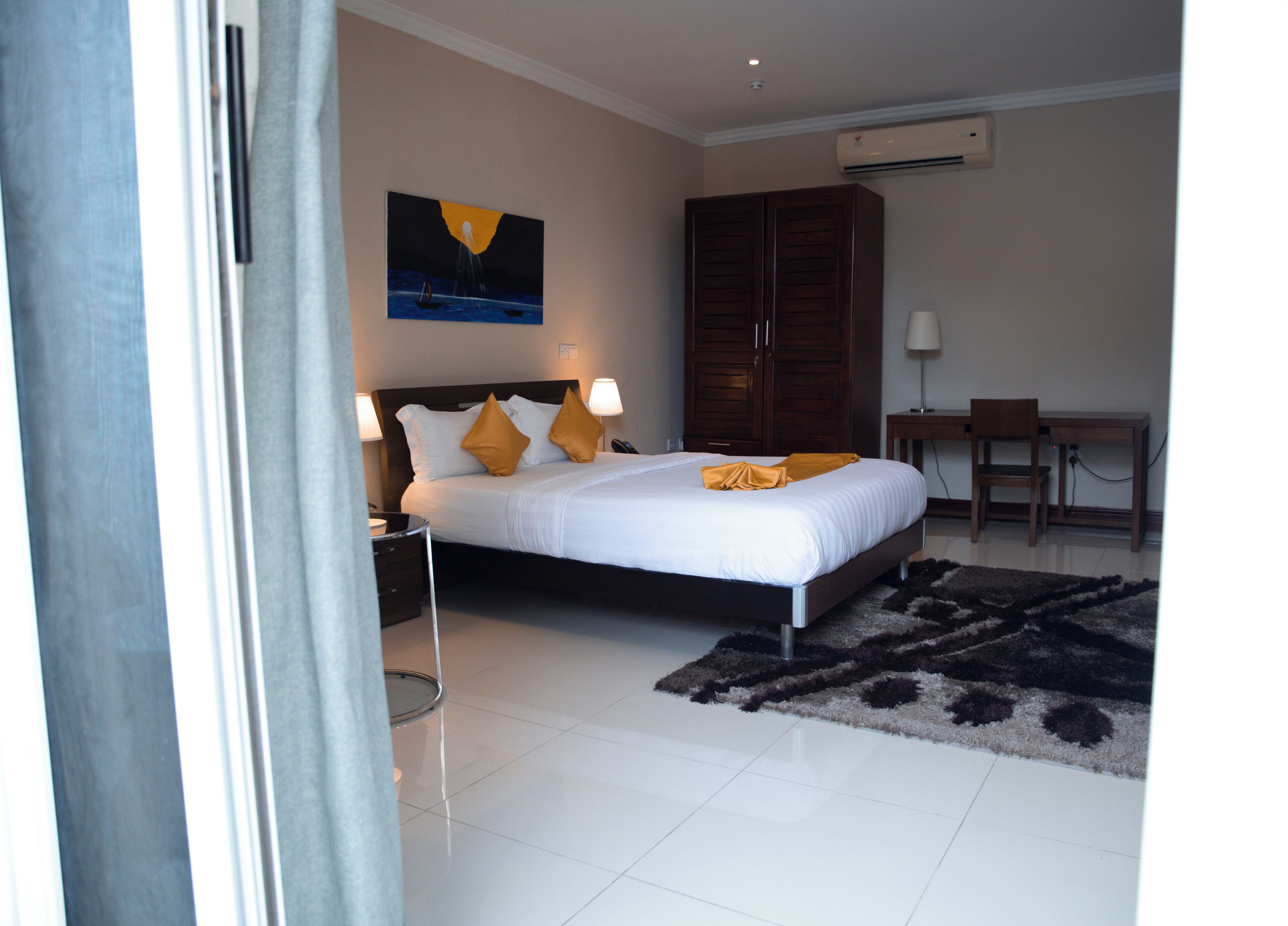 A hotel room with a bed, a desk, a chair, a painting on the wall, and an air conditioning unit.