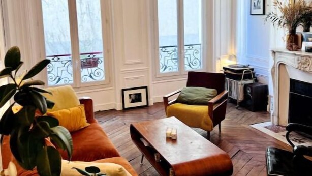 Amplia sala de estar con piso de madera, un ventanal con vistas, chimenea y sofá con cojines.