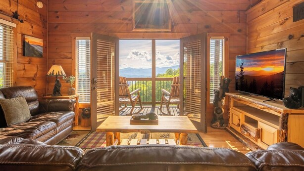 A wooden cabin interior with a leather sofa, a table with a tray, a TV, and a balcony with a mountain view.