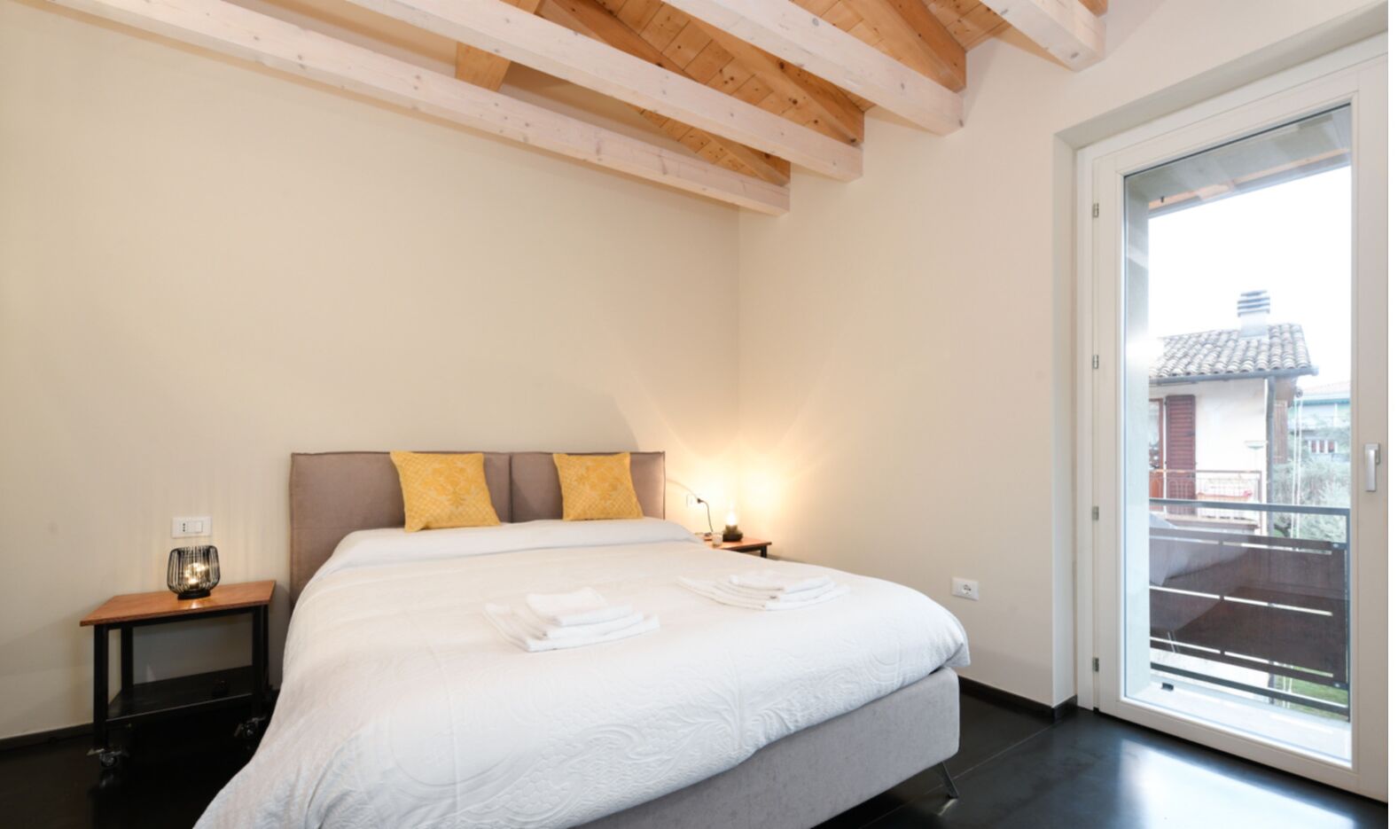 A bedroom with a large bed, wooden beams on the ceiling, a nightstand with a lamp, and a view of a building through a glass door.