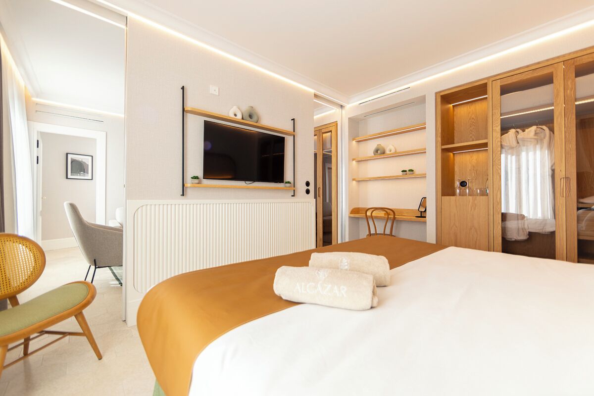 Una habitación de hotel con cama, televisión montada en la pared, armario de madera, mesita pequeña y silla.