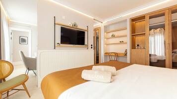 A hotel room with a bed, a TV mounted on the wall, a wooden wardrobe, a small table, and a chair.