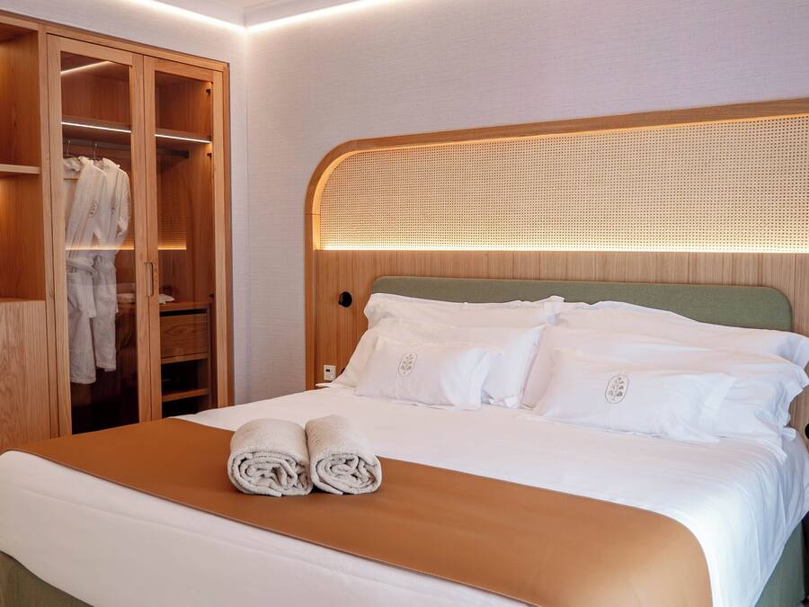 Una cama bien hecha con sábanas blancas y dos toallas enrolladas, un armario de madera con puertas de cristal, y un taburete de madera.