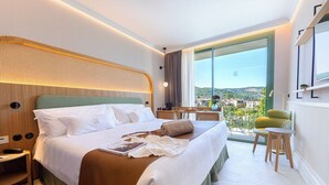 Ein modernes Hotelzimmer mit einem großen Bett, einem kleinen Schreibtisch, einem Sessel und Blick auf die Stadt.