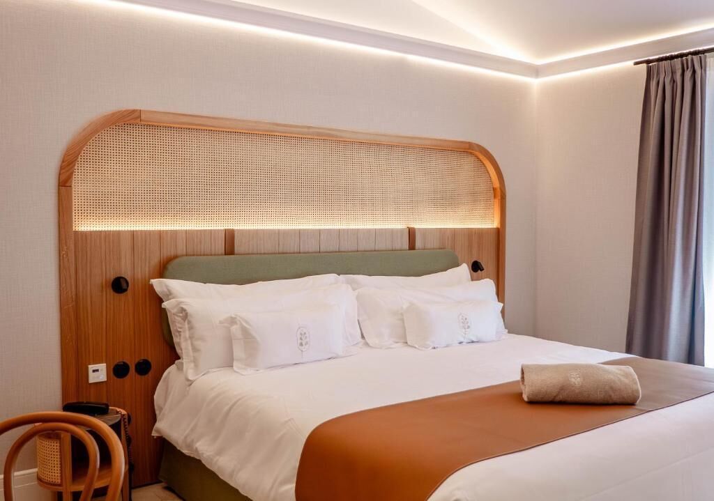 Una cama bien hecha con sábanas blancas y una manta marrón, un cabecero de madera con panel trasero tejido, y una silla de madera con asiento tejido.