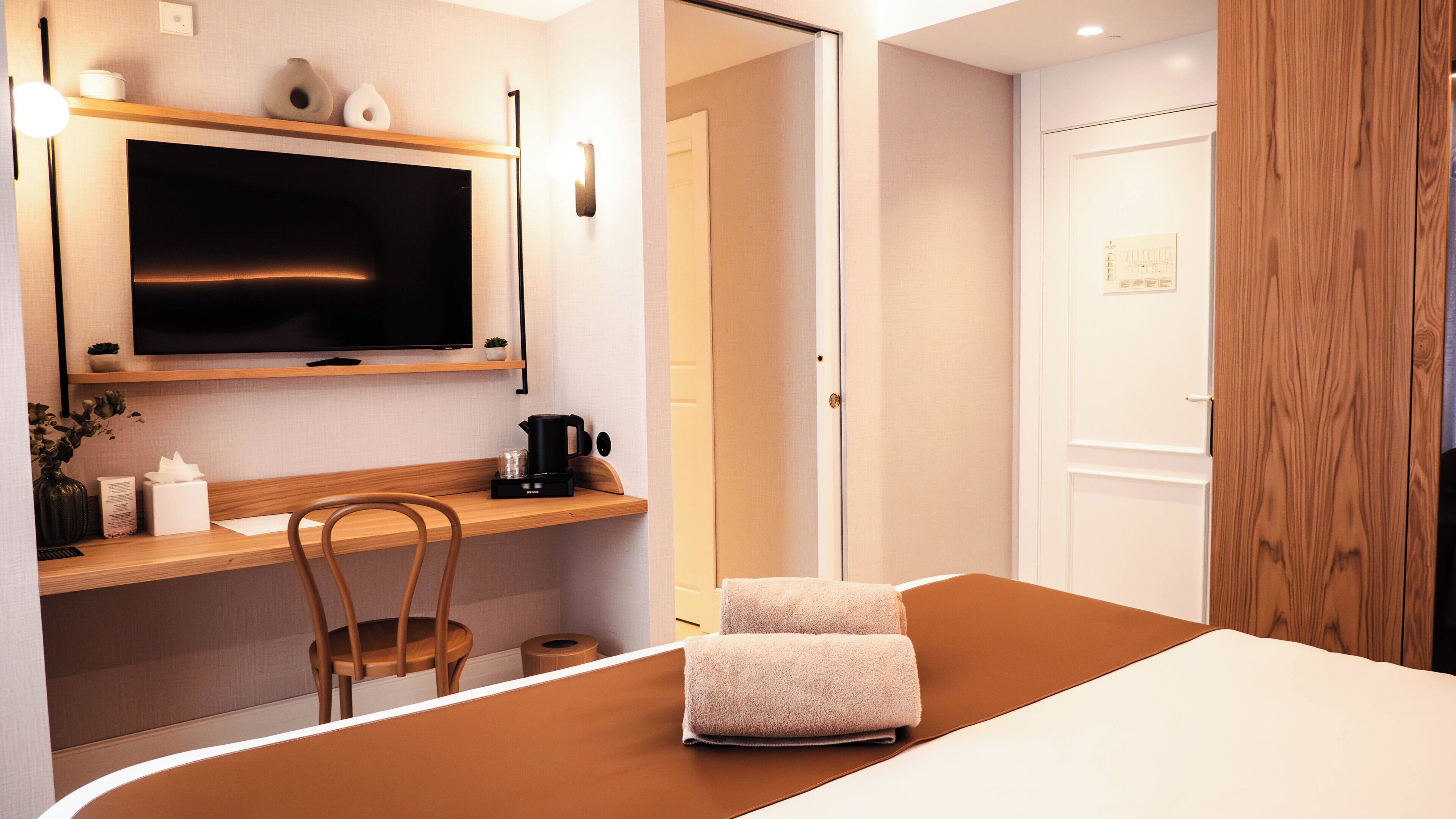 A hotel room with a bed, a desk with a chair, a television, a coffee maker, and a door.