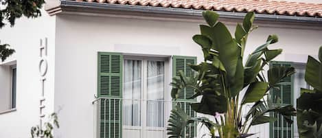 A hotel facade with a tiled roof, green shutters, and a balcony with plants.