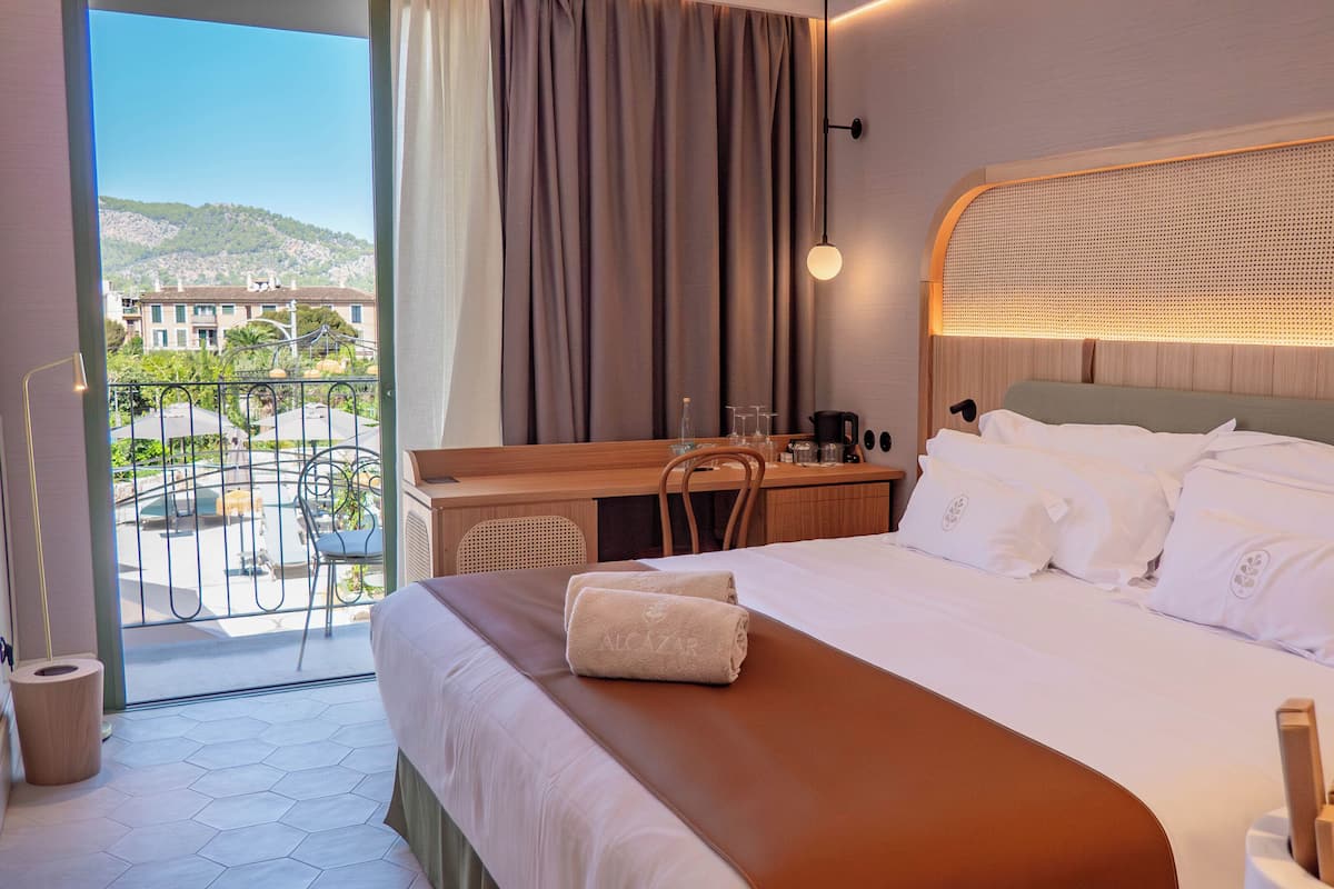 A hotel room with a bed, a desk, and a view of a balcony with outdoor furniture.