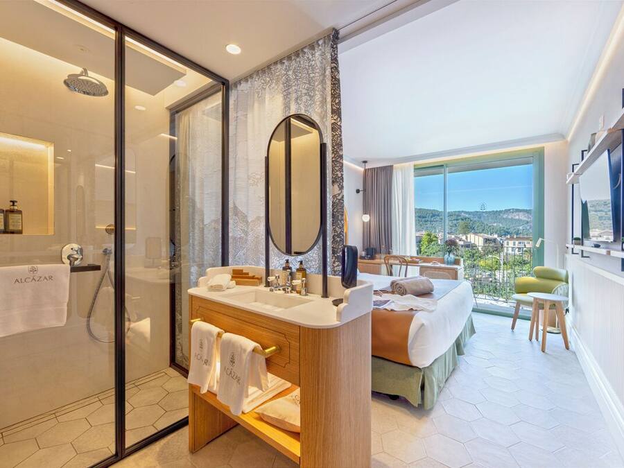 Une chambre d’hôtel moderne dotée d’une douche en verre, d’un grand miroir, d’une coiffeuse en bois, d’un lit recouvert d’une courtepointe verte et offrant une vue sur la ville grâce à de grandes fenêtres.