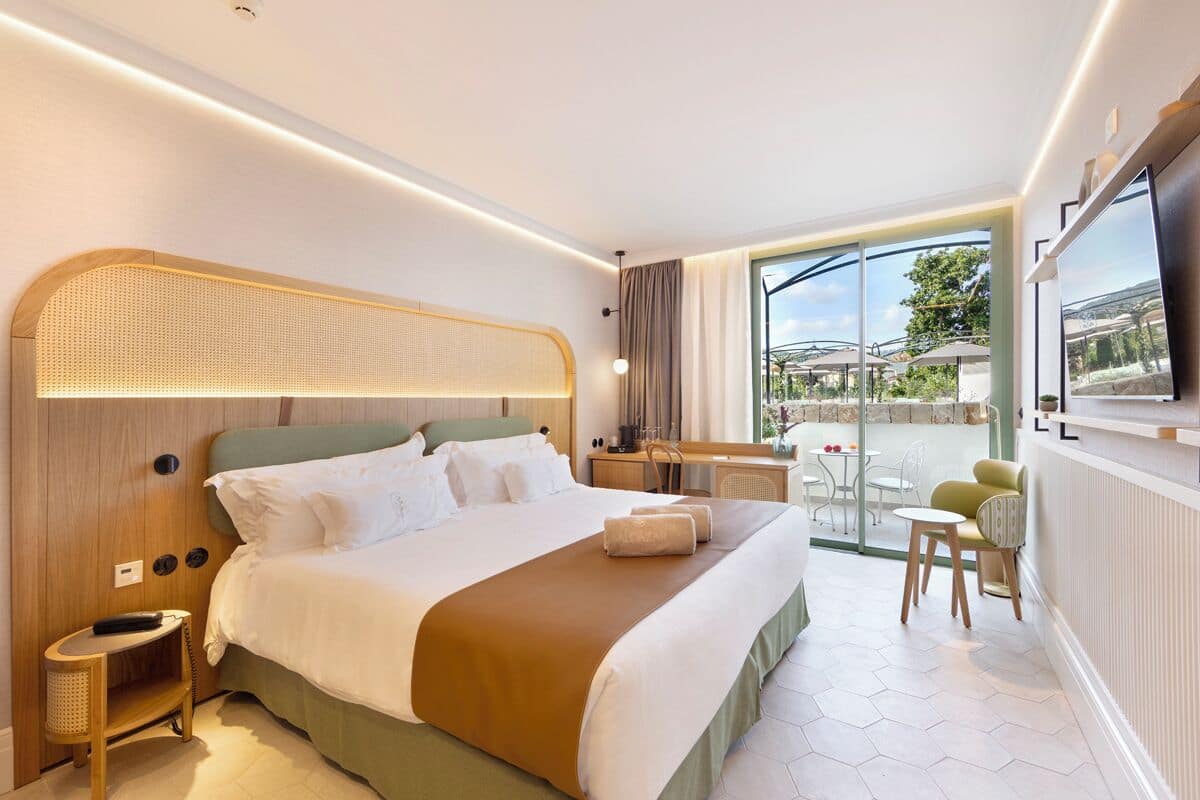A modern hotel room with a large bed, a desk, and a balcony with outdoor seating.