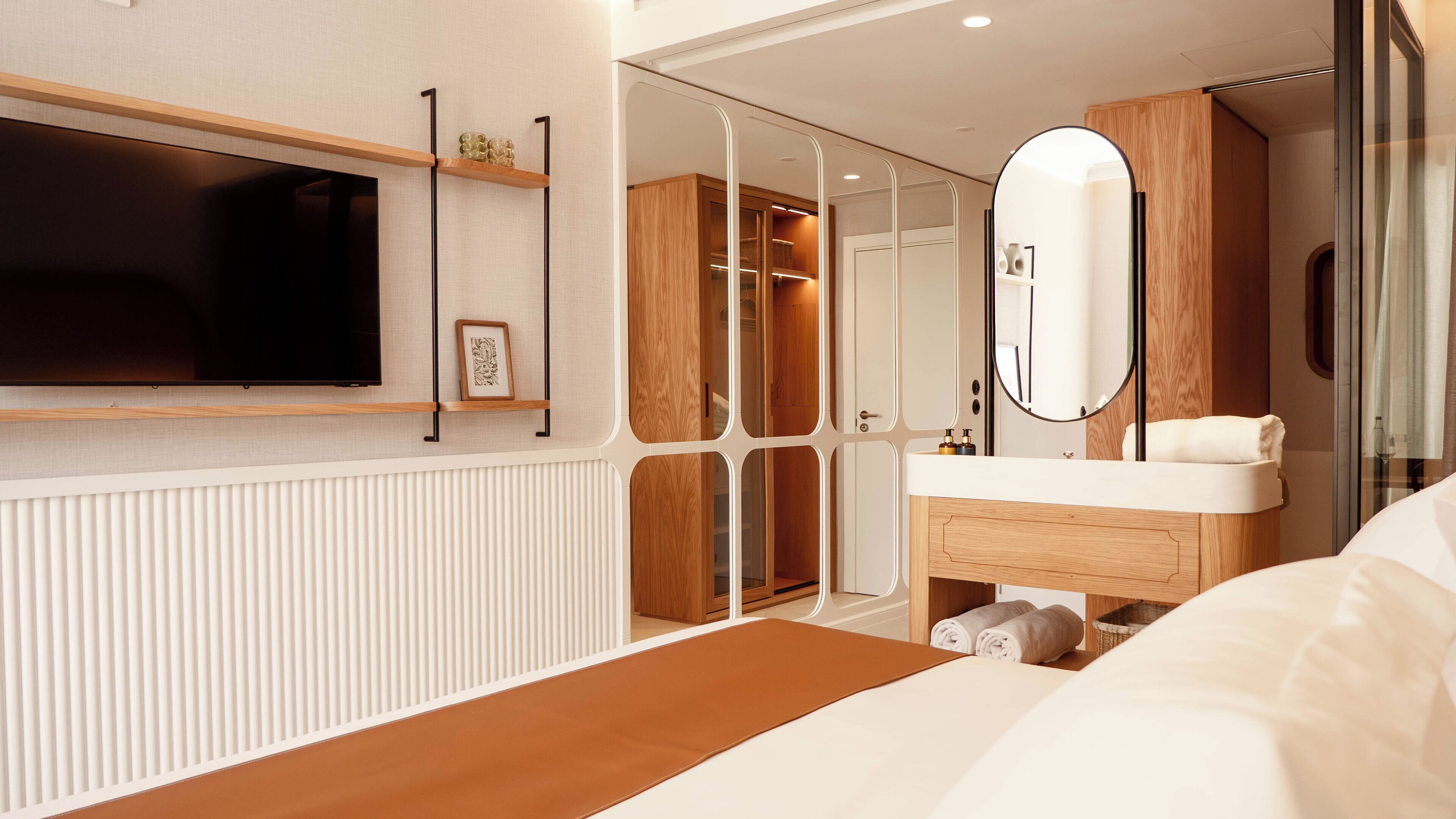 A modern hotel room with a large bed, a TV mounted on the wall, a wooden vanity with a mirror, and a walk-in closet.