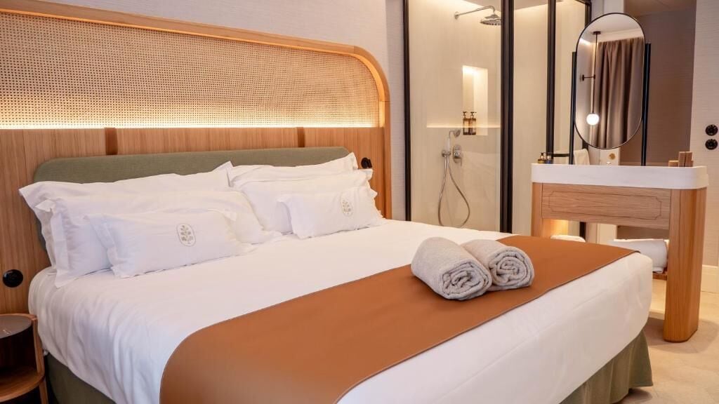 A hotel room with a large bed, a wooden headboard, a bathroom with a mirror, and a towel on the bed.
