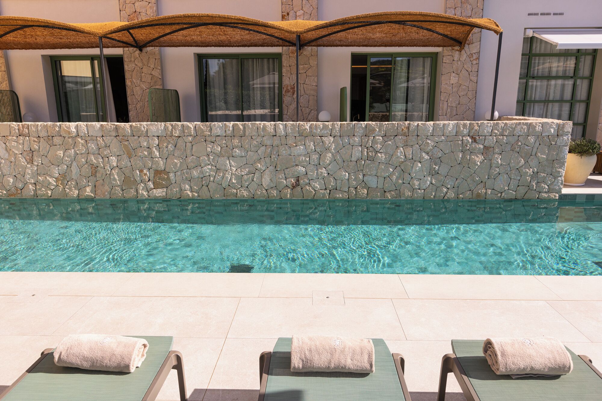 A swimming pool with clear water, surrounded by lounge chairs with towels, and a stone wall with windows in the background.