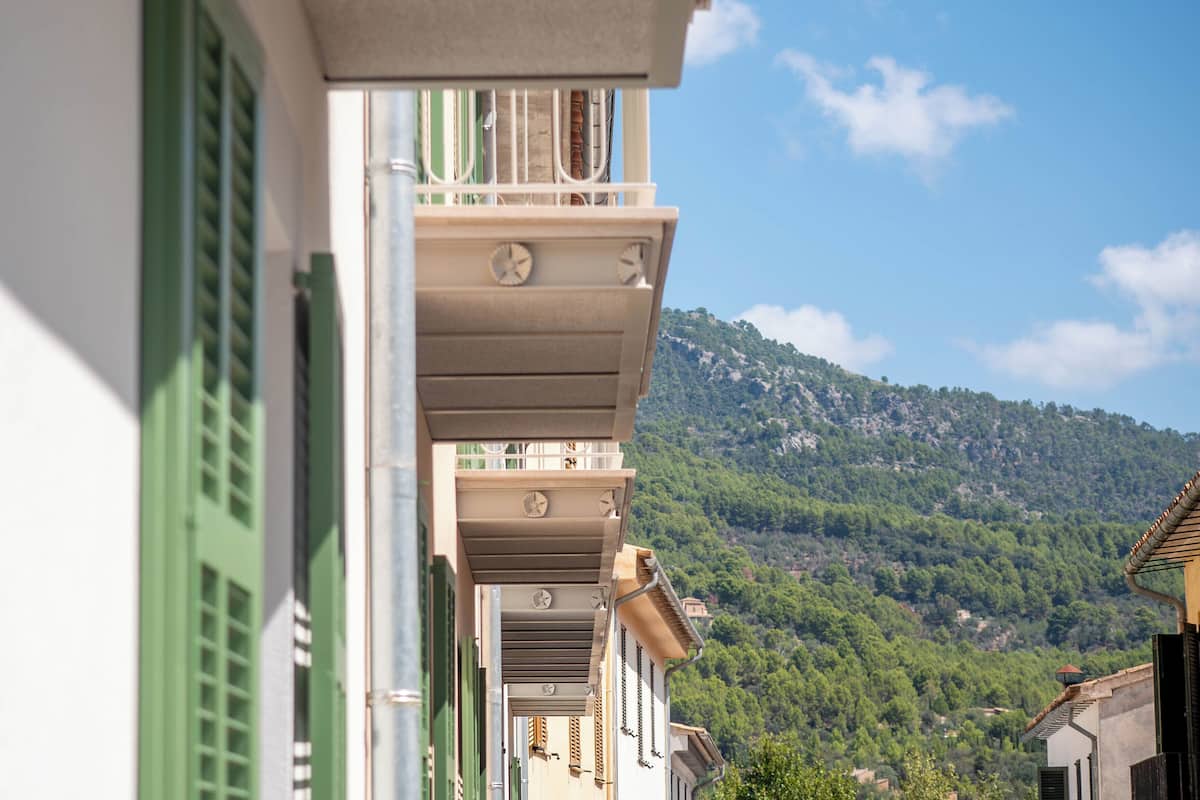 A narrow street with buildings featuring balconies and green shutters, leading to a hillside with dense vegetation.