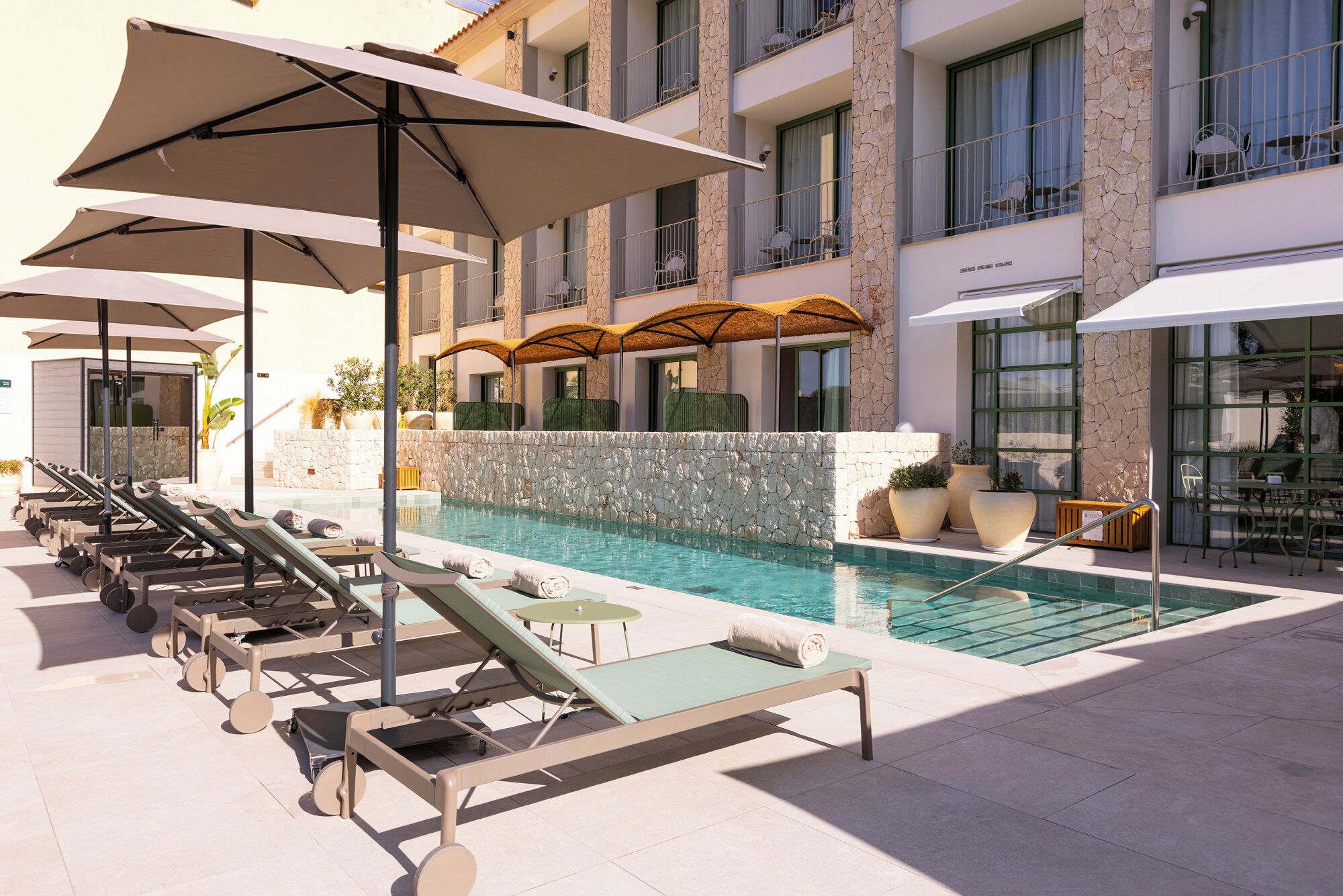 Un área junto a la piscina con sillones, sombrillas y un muro de piedra.