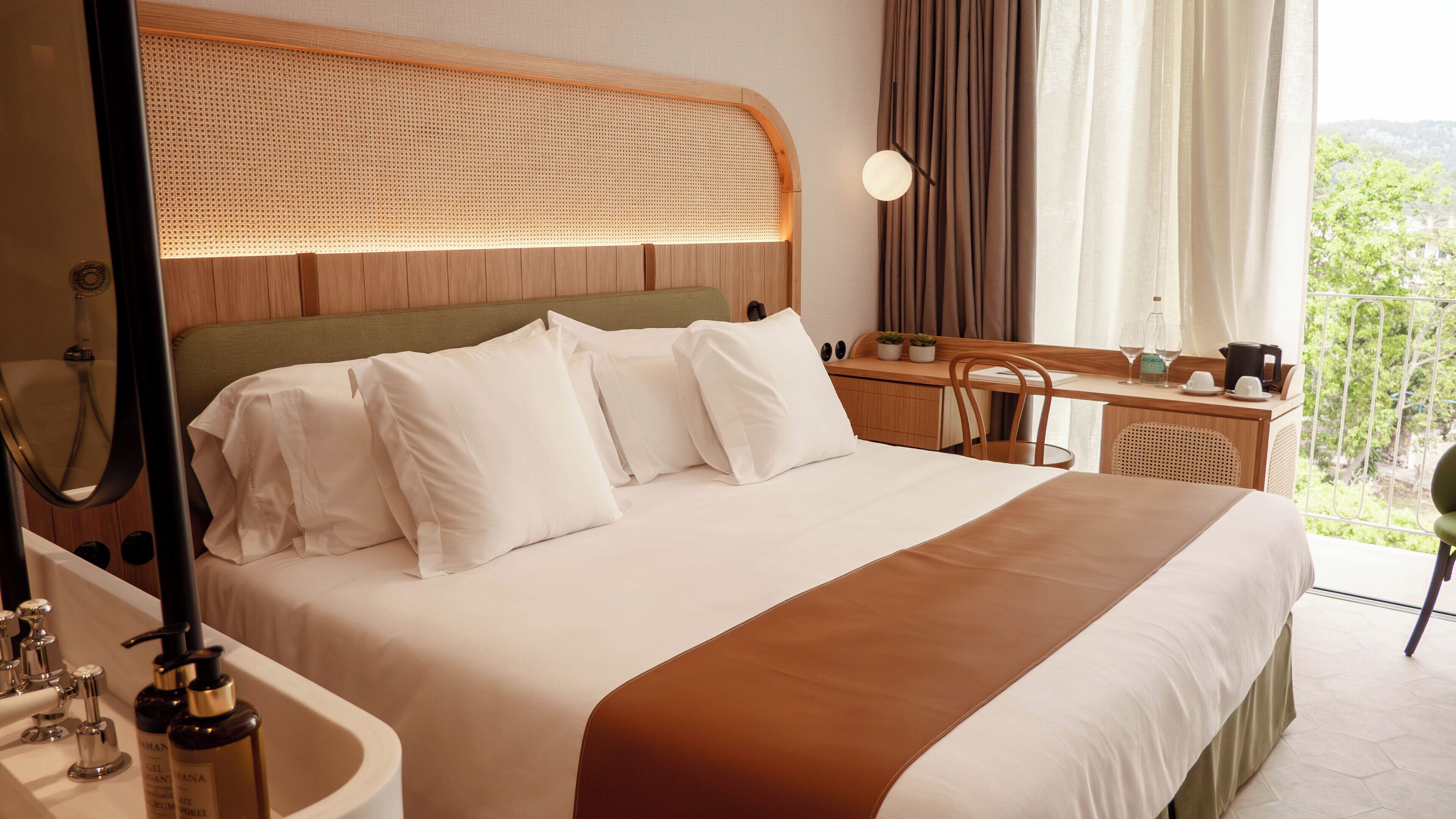 A hotel room with a large bed, a desk, a chair, a window with curtains, and a view of greenery.