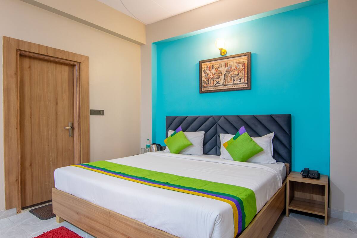 Un dormitorio con puerta de madera, cama con colcha blanca y colorida, pared de acento azul y una mesita de noche pequeña con un teléfono.