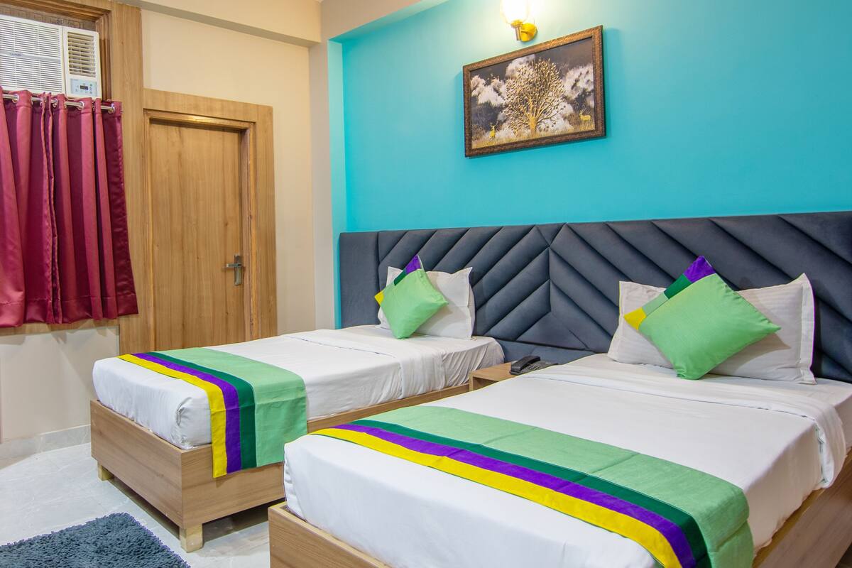 Habitación de hotel con dos camas, cada una con colcha a rayas, cabecero y almohadas. Se observa una ventana con cortinas, una lámpara fijada a la pared y un cuadro enmarcado sobre las camas.