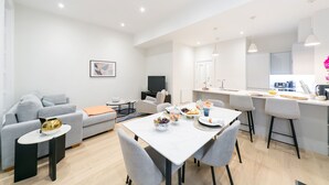 A modern living room with a grey sofa, a small round table, and a TV. There is a dining area with a white table and grey chairs. The kitchen has a white countertop and a sink. - Urban Chic - Acre (London)