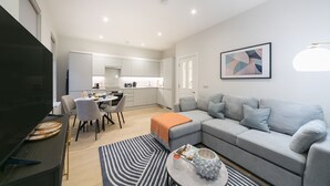 A modern living room with a grey sofa, a dining table with chairs, a TV, and a kitchen in the background. - Urban Chic - Acre (London)