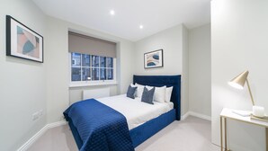 A bedroom with a bed, a window, a bedside table with a lamp, and two framed pictures on the wall. - Urban Chic - Acre (London)