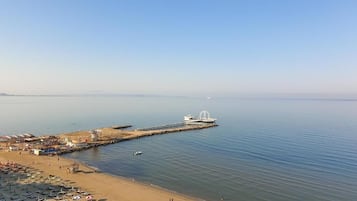 Une plage bordée de rangées de parasols et de chaises longues, une jetée surmontée d’une structure blanche, et une eau calme.