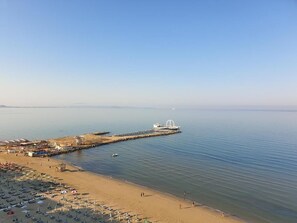Una playa con hileras de sombrillas y tumbonas, un muelle con una estructura blanca y agua tranquila.