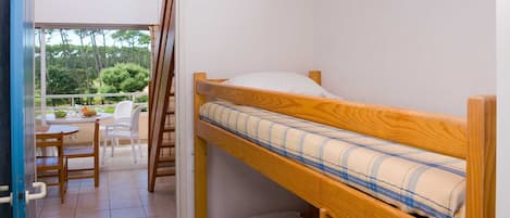 Une petite chambre avec un seul lit, un lit superposé en bois, une petite table avec des chaises et une vue sur un jardin par une porte ouverte.