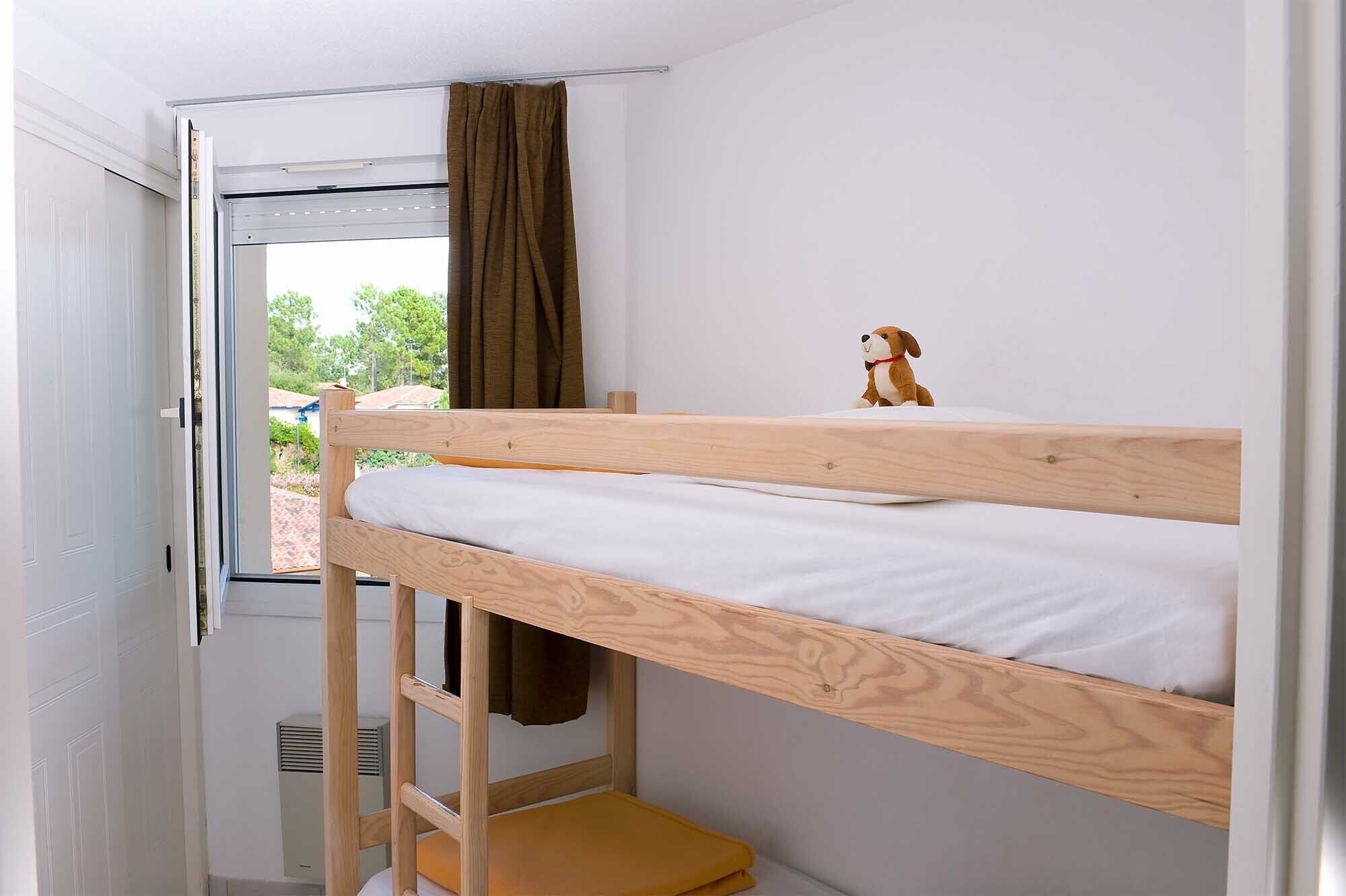 Un lit superposé avec une structure en bois et une literie blanche, un petit jouet posé sur le lit du haut, et une fenêtre avec un rideau marron.