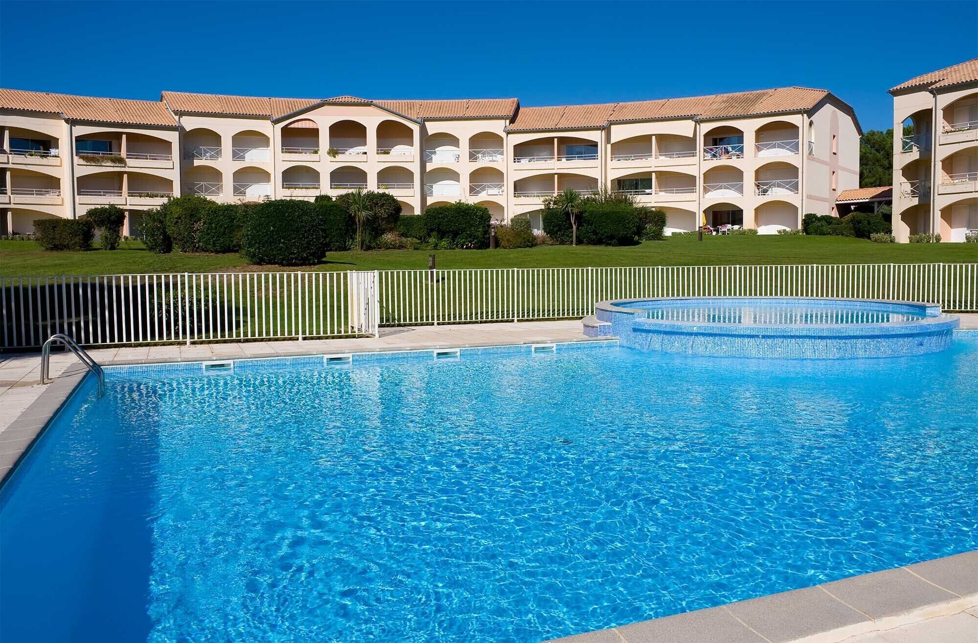 A swimming pool with a clear blue water surface, surrounded by a white fence. In the background, there are multi-story buildings with balconies and a well-maintained lawn.